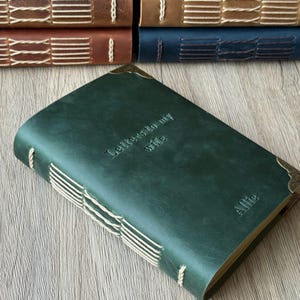 以下が含まれることがあります： 「Letters to my wife」のエンボス加工が施された、名前「Allie」入りの濃い緑色の革製ジャーナル。背表紙に沿ってクリーム色のステッチが施され、真鍮製のコーナープロテクターが付いています。他のジャーナルが背景に見えます。