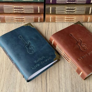 May include: A blue and a brown leather journal with a guitar and musical note design embossed on the cover. The blue journal has the text "Emily's Lyric Journal". The journals have stitched binding and brass corner protectors. A stack of similar journals is in the background.