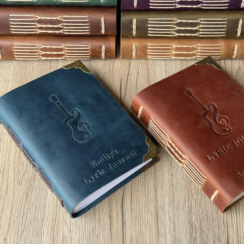 May include: A blue and a brown leather journal with a guitar and musical note design embossed on the cover. The blue journal has the text "Emily's Lyric Journal". The journals have stitched binding and brass corner protectors. A stack of similar journals is in the background.
