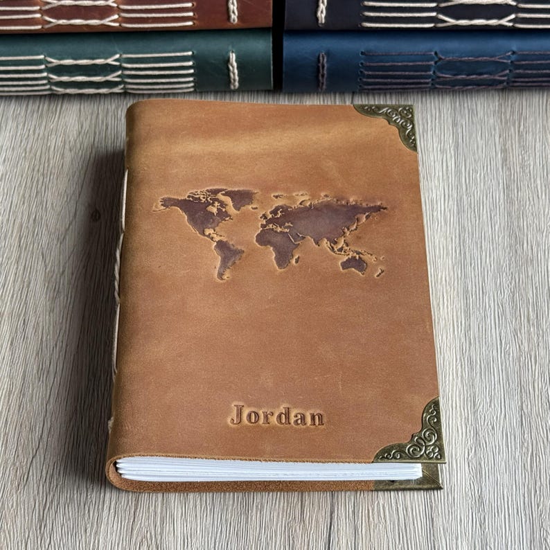 May include: A brown leather journal featuring a world map design embossed on the cover. The name "Jordan" is stamped below the map. The journal has decorative corner pieces and a visible binding.