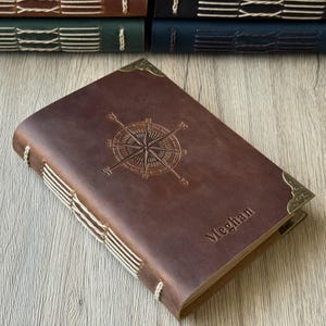 Könnte beinhalten: Ein braunes Lederjournal mit einem Kompassrosen-Design und dem Namen "Meghan" auf dem Einband. Das Journal hat dekorative Eckstücke und ist mit weißen Fäden gebunden. Weitere Journale sind im Hintergrund zu sehen.