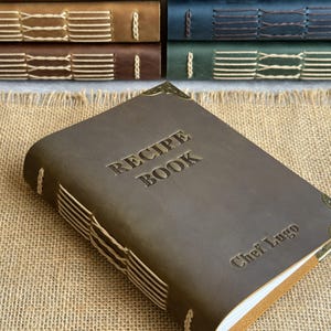 Może przedstawiać: Brązowa sk&oacute;rzana książka kucharska z wytłoczonymi słowami "RECIPE BOOK" na okładce. Książka ma ozdobne przeszycia wzdłuż grzbietu i ochraniacze narożnik&oacute;w. Imię "Chef Lugo" jest r&oacute;wnież wytłoczone na okładce. W tle widocznych jest kilka innych książek.