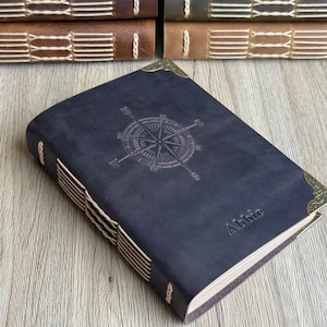 May include: A dark blue leather journal featuring an embossed compass rose design. The journal has decorative corner pieces and is hand-stitched with cream thread. The name "Abbie" is embossed on the cover. Other journals are visible in the background.