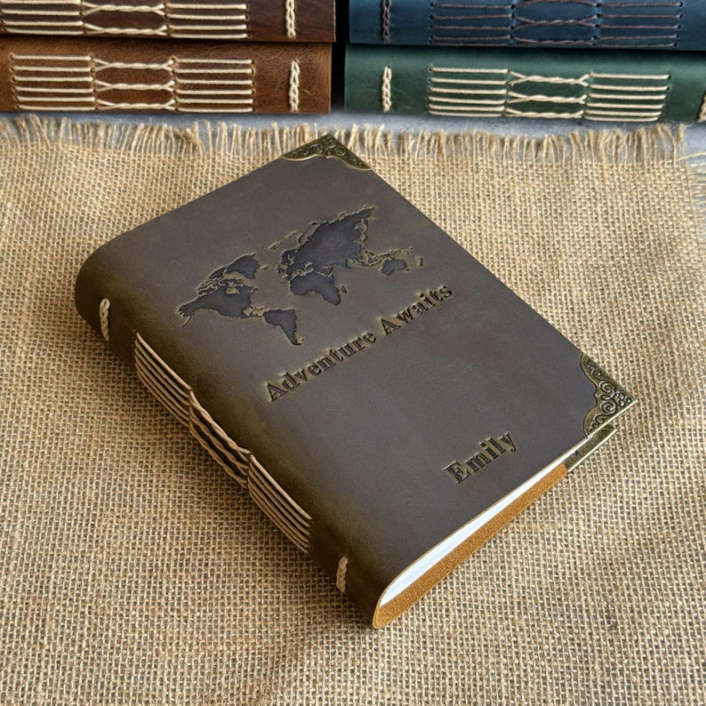 May include: A brown leather journal with the words "Adventure Awaits" and "Emily" embossed on the cover. The journal features a world map design and decorative corner accents. The pages are bound with white stitching.