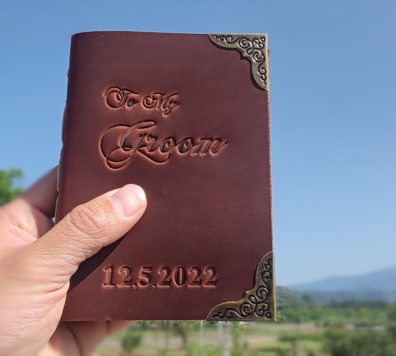 May include: A brown leather journal with a decorative brass corner piece. The journal has the words "To My Groom" embossed on the front cover and the date "12.5.2022" embossed below.