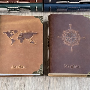 May include: Two leather journals, one tan with a world map, the other brown with a compass rose. Both have decorative corner accents and embossed names: "Jordan" and "Meghan". The journals are suitable for travel or personal use.