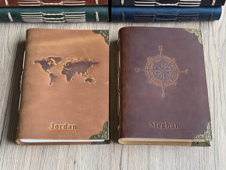 May include: Two leather journals, one tan with a world map, the other brown with a compass rose. Both have decorative corner accents and embossed names: "Jordan" and "Meghan". The journals are suitable for travel or personal use.