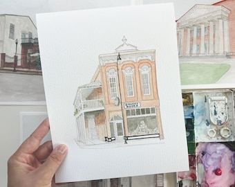Op de afbeelding: Aquarel schilderij van een gebouw met de tekst "Square Books" boven de winkelpui. Het gebouw is perzik van kleur met witte randen en een balkon. Andere aquarel schilderijen staan op de achtergrond.