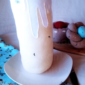 May include: A white felt Easter cake with colorful sprinkles on top, sitting on a white plate. The cake is on a blue and white patterned tablecloth, next to a brown felt pastry.