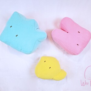 May include: Three plush toys in the shape of cartoon characters. The blue character is a star shape, the pink character is a bunny shape, and the yellow character is a fish shape. The toys are on a white background.