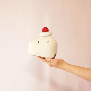 Puede incluir: Un juguete de peluche crema, de forma rectangular, que se asemeja a un pastel, con una pequeña esfera roja en la parte superior. El juguete tiene dos pequeños ojos negros y una base marrón. Se sostiene en la mano de una persona sobre un fondo rosa.