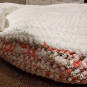 May include: A white and peach colored round pet bed with a fluffy texture. The bed is made of yarn and has a soft, cozy appearance.