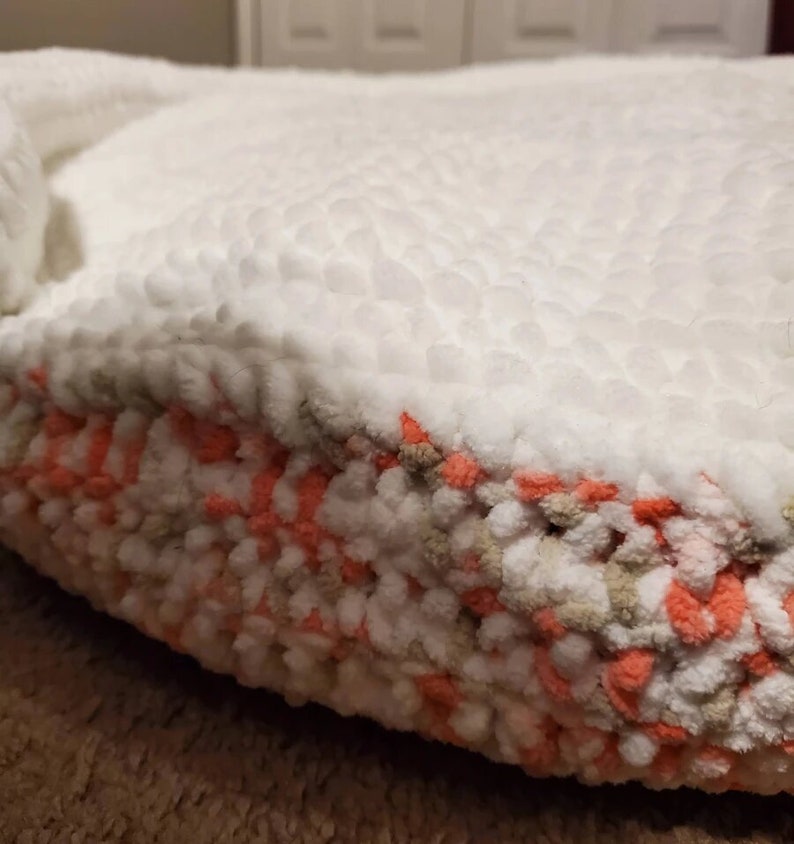May include: A white and peach colored round pet bed with a fluffy texture. The bed is made of yarn and has a soft, cozy appearance.