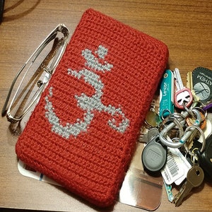 Può includere: Borsa rossa all'uncinetto con un design a forma di ancora bianca. La borsa è appoggiata su una superficie di legno con un set di chiavi e un astuccio per occhiali.