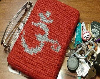 May include: Red crocheted pouch with a white anchor design. The pouch is lying on a wooden surface with a set of keys and a glasses case.