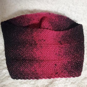 May include: A black and pink ombre knitted neck warmer. The neck warmer is made of a soft, textured yarn.