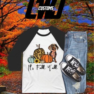 Könnte beinhalten: Ein weißes und schwarzes Raglan-Ärmel-Shirt mit einem Dackel- und Kürbis-Motiv. Das Shirt wird mit einer blauen Jeans und schwarzen Turnschuhen kombiniert. Der Text "It's Fall Y'all" ist auf das Shirt gedruckt.