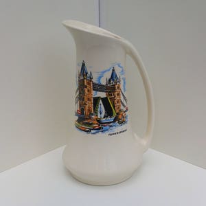 May include: White ceramic jug with a colourful image of Tower Bridge in London, England. The jug has a handle and the text "Tower Bridge" is printed on the bottom.