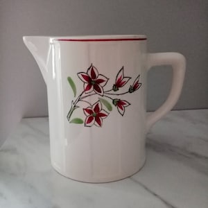 Op de afbeelding: Witte keramische kan met een rode rand en een bloemenontwerp met rode en groene bloemen. De kan heeft een handvat en een tuit.