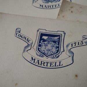 Op de afbeelding: Close-up van een vintage papier met een blauw en wit embleem. Het embleem toont een schild met een vogel en andere symbolen, met de woorden "COGNAC" en "MARTELL" op een banner. Het jaar "1715" is ook zichtbaar.