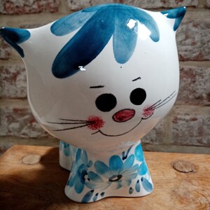 Può includere: Una statuetta di gatto in ceramica bianca con disegni floreali blu e bianchi. Il gatto ha un design dipinto blu e bianco sulla testa e un naso rosa.