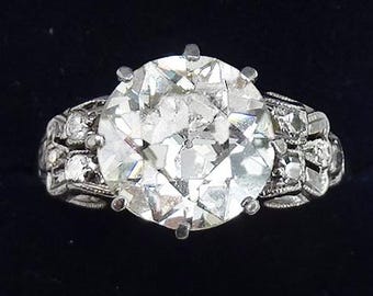 Puede incluir: Un anillo de plata con un gran diamante redondo y claro en el centro. El diamante está rodeado de diamantes más pequeños engastados en una banda de plata.