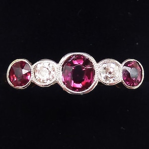 Peut inclure: Une bague en argent sertie de cinq pierres rondes. La pierre centrale est un gros rubis rouge foncé. Les deux pierres de chaque côté du rubis sont des diamants blancs ronds plus petits. Les deux pierres extérieures sont des rubis rouges foncés ronds plus petits.