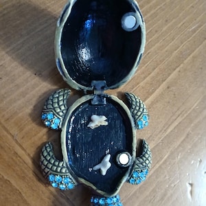 May include: A decorative turtle-shaped trinket box with an open lid. The box is black and gold with blue gemstone accents. The interior is black with two white decorative pieces and a magnetic closure.