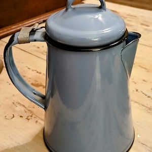 May include: A vintage blue enamel coffee pot with a black rim and handle. The pot has a lid and a spout for pouring. The handle is attached to the side of the pot with a metal bracket. The pot is sitting on a light-colored wooden surface.
