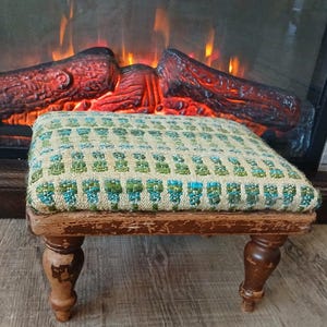 May include: An antique footstool with a rectangular, patterned cushion in shades of green and blue. The wooden frame shows signs of wear, with four turned legs. The background features a fireplace with faux logs and flickering lights.