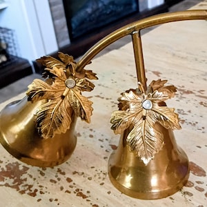 May include: A gold-toned metal double bell ornament featuring two bells suspended from a curved bar. Each bell is adorned with a textured metallic maple leaf accent at the top, centered with a small, clear, circular stone detail.