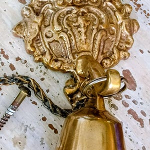 May include: A brass bell attached to a decorative wall-mounted bracket with an ornate, embossed design. The bell hangs from a metal ring connected to a short chain, and a metal screw with a threaded end is attached to the side of the bracket.