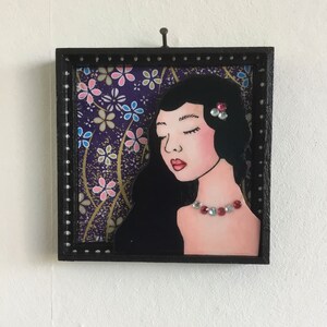 Op de afbeelding: Een vierkante zwarte lijst met een mixed-media kunstwerk van een persoon met lang zwart haar, gesloten ogen en roze lippen. De figuur draagt een ketting en een haaraccessoire gemaakt van kleine, kleurrijke strass-steentjes tegen een paarse bloemenachtergrond.