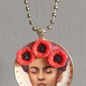 Può includere: Una collana con ciondolo rotondo color argento raffigurante il ritratto di una persona con capelli scuri e tre fiori di papavero rossi tridimensionali con centri neri sulla parte superiore. Il ciondolo pende da una catena a palline argentata.