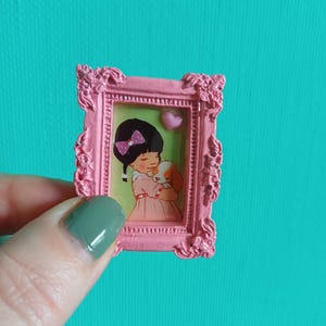 Può includere: Una piccola cornice rosa ornata con un'illustrazione a cartoni animati di una ragazza in un vestito rosa che tiene un cucciolo. La cornice ha un dettaglio a forma di cuore e uno sfondo turchese.