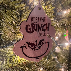 Puede incluir: Un adorno navideño de madera con forma de la cara del Grinch con el texto "Resting Grinch Face" en él.