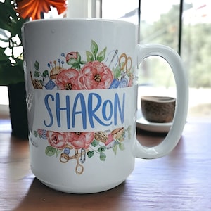 May include: White ceramic mug with a floral design and the name "SHARON" in blue lettering. The design includes sewing-themed imagery like spools of thread, buttons, and scissors.