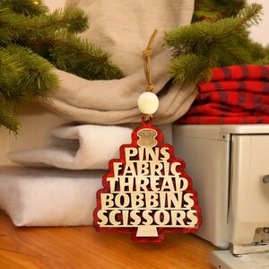 May include: Red wooden Christmas tree ornament with the words "Pins, Fabric, Thread, Bobbins, Scissors" written on it.