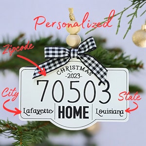 Puede incluir: Adorno navideño blanco con un lazo a cuadros blanco y negro. El adorno tiene forma de letrero con el texto "CHRISTMAS -2023- 70503 Lafayette HOME Louisiana" y la palabra "Personalized" en la parte superior.