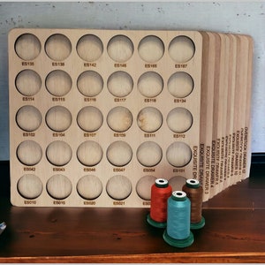 May include: A wooden storage box with 24 circular compartments labeled with numbers. The box is filled with spools of thread in red, blue, and brown.