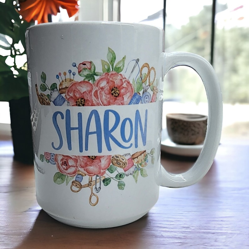 May include: White ceramic mug with a floral design and the name "SHARON" in blue lettering. The design includes sewing-themed imagery like spools of thread, buttons, and scissors.