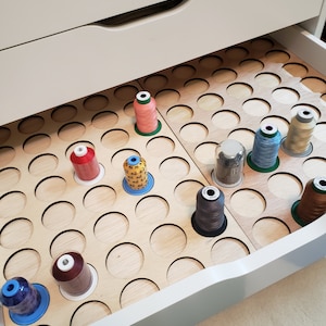 May include: A white drawer with a wooden organizer for storing spools of thread. The organizer has many circular cutouts to hold the spools. There are several spools of thread in various colors, including red, orange, yellow, brown, blue, and green.