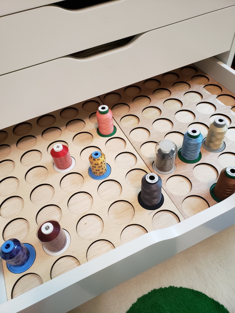 May include: A white drawer with a wooden organizer for storing spools of thread. The organizer has many circular cutouts to hold the spools. There are several spools of thread in various colors, including red, orange, yellow, brown, blue, and green.