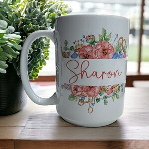 May include: White ceramic mug with a floral design featuring pink flowers, green leaves, and sewing-themed accents. The name "Sharon" is written in a cursive font on a banner.