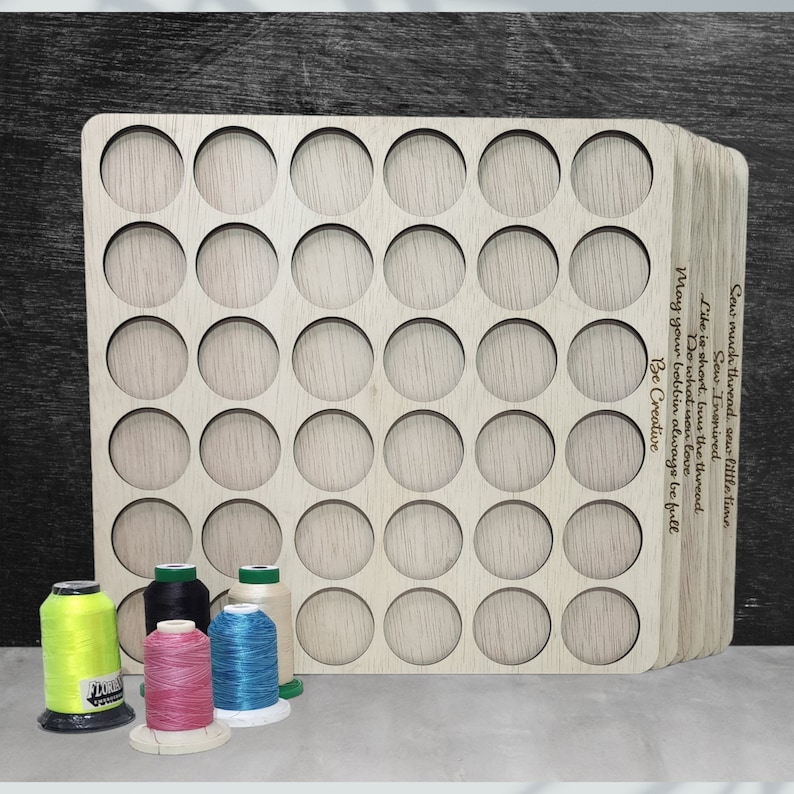 May include: A wooden thread organizer with 36 circular slots for storing spools of thread. The organizer is made of light wood and has a natural finish. The text "Sew much thread sew little time. Sew Inspired. Like is short, but the thread. Do what you love. May your bobbin always be full. Be Creative" is engraved on the side of the organizer.