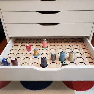 May include: A white drawer with a wooden organizer for storing spools of thread. The organizer has multiple circular slots and is divided into two sections. The drawer is open and there are several spools of thread in various colors, including red, blue, green, and brown.