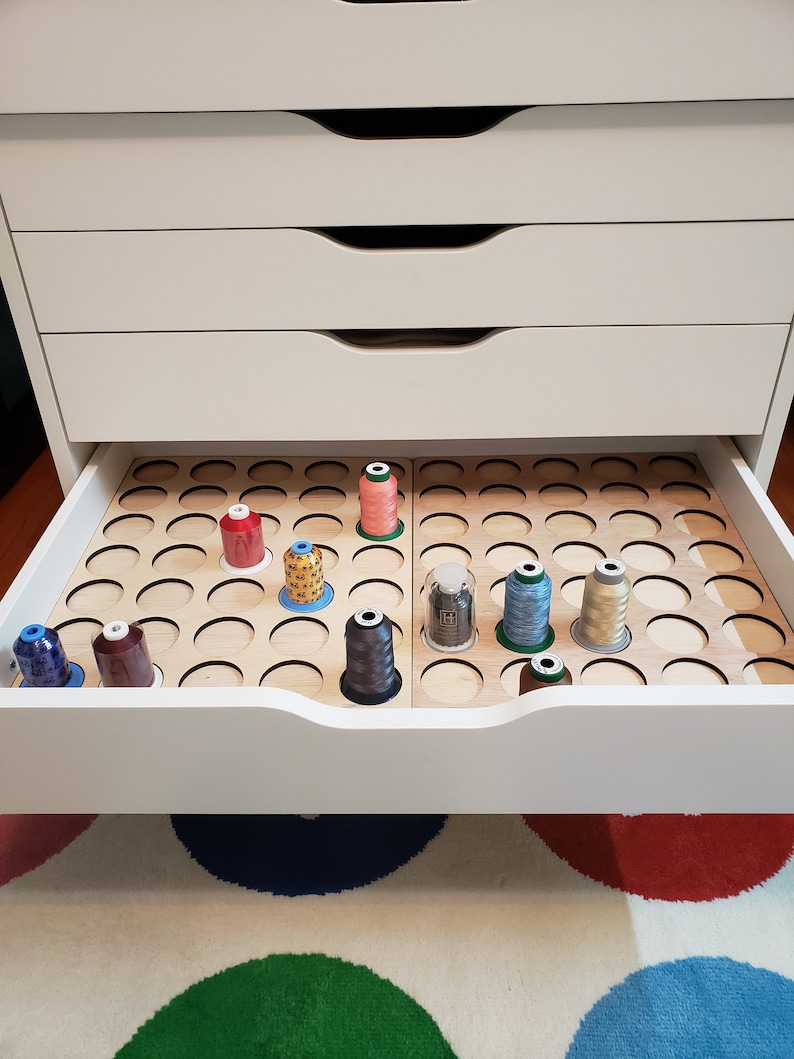 May include: A white drawer with a wooden organizer for storing spools of thread. The organizer has multiple circular slots and is divided into two sections. The drawer is open and there are several spools of thread in various colors, including red, blue, green, and brown.