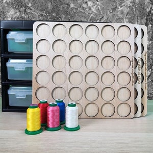 May include: A wooden thread organizer with 36 circular slots for storing spools of thread. The organizer has the text "One stitch at a time" and "Happiness is a full bobbin" engraved on it. Five spools of thread in different colors are in the foreground.