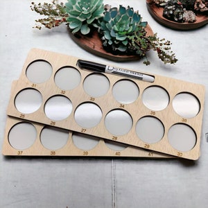 May include: A wooden paint palette with 41 circular wells for holding paint. The palette is made of light wood and has a natural finish. The wells are numbered from 25 to 41. A black dry erase marker is lying on top of the palette.