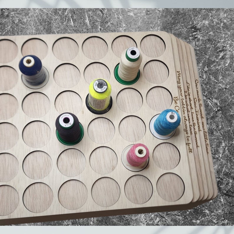 May include: A wooden thread organizer with 49 circular slots for storing spools of thread. The organizer has a quote engraved on the side: "Cow much thread sew little time. Sew Inspired. Life is short but the thread Do what you love. May your bobbin always be full. Be Cr"